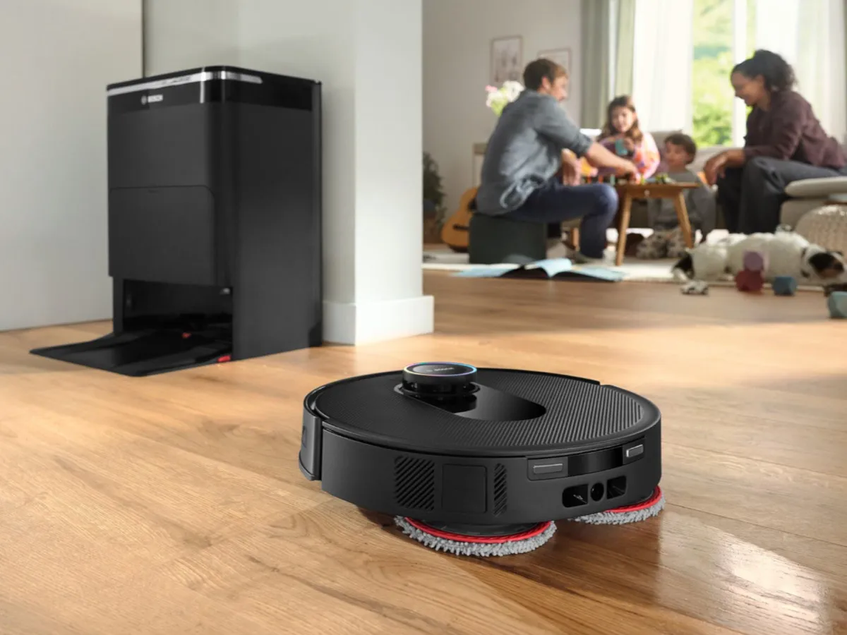 Un robot aspirapolvere Bosch passa su un pavimento in legno chiaro e pulisce accuratamente il pavimento, mentre sullo sfondo una famiglia è seduta a tavola e trascorre del tempo insieme.