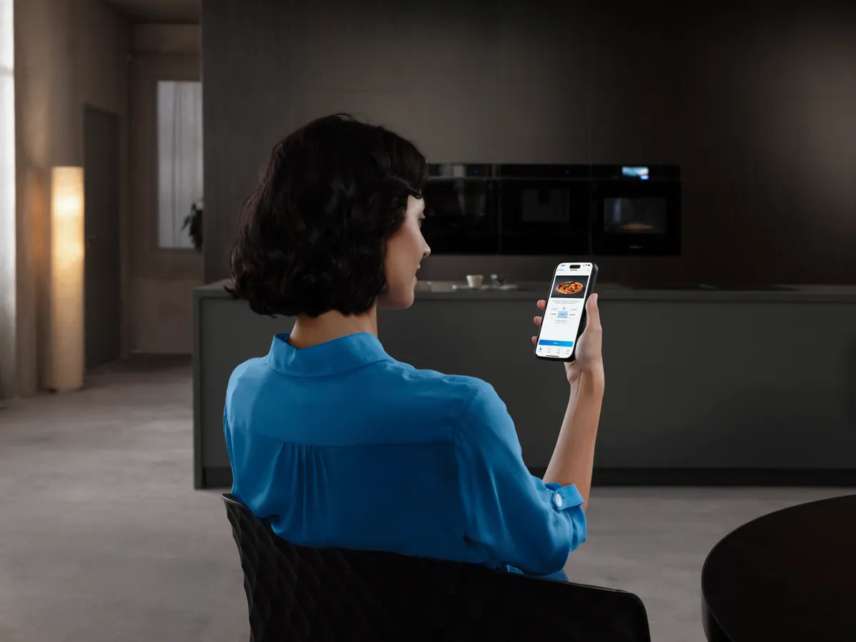 Una persona con una maglietta blu tiene in mano uno smartphone su cui è visualizzata un'app per la consegna di cibo a domicilio, seduta in una cucina moderna.