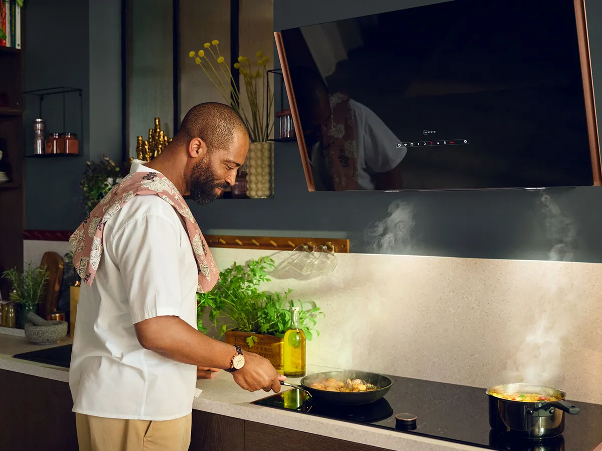 Un homme se tient devant une plaque de cuisson, préparant des aliments dans une poêle et une casserole, de la vapeur s'échappant de chacune.