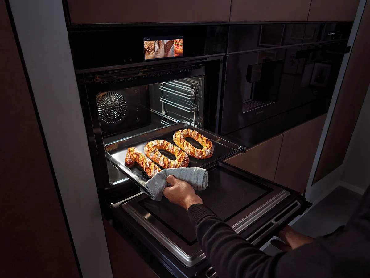 A person is pulling out a tray of freshly baked pretzels from a modern oven, showcasing a sleek kitchen interior.