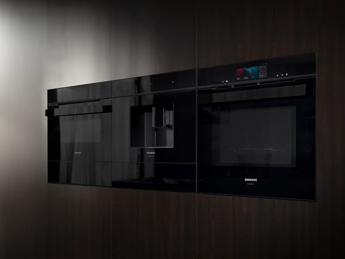 Three sleek black kitchen appliances by Siemens, integrated into a dark wood panel, showcasing modern design and technology.