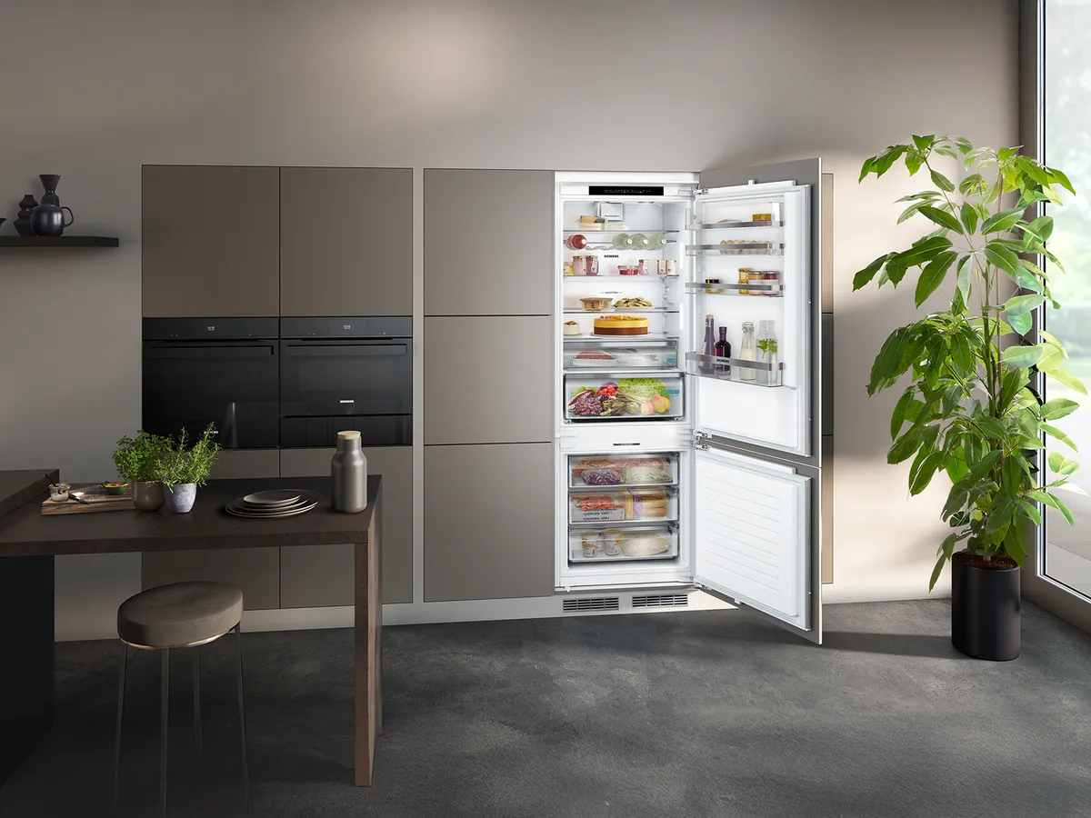 A modern kitchen featuring a stylish, open refrigerator filled with food, alongside a dining table and a green indoor plant.
