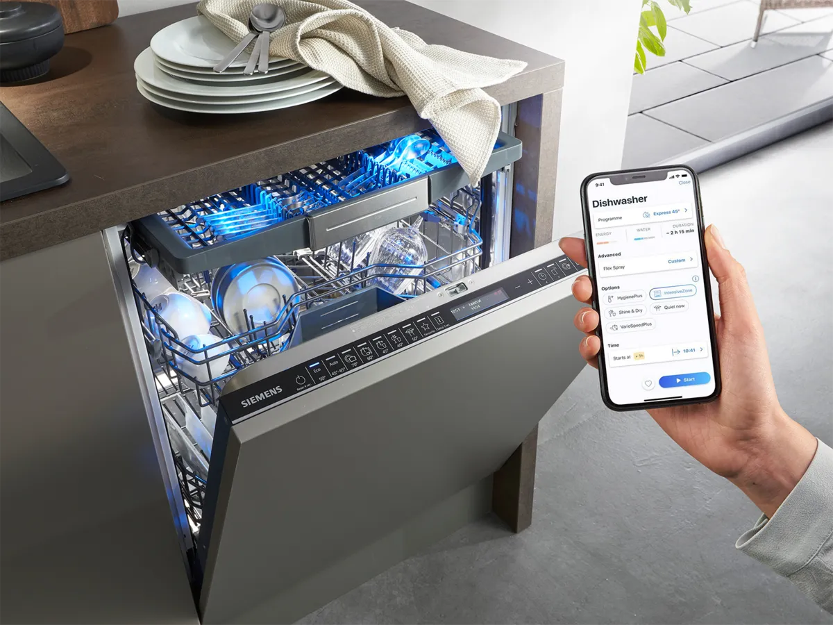 Ręka trzyma smartfon z aplikacją Siemens, obok stoi otwarta zmywarka z talerzami i podświetlonym wnętrzem, prezentująca inteligentne funkcje.