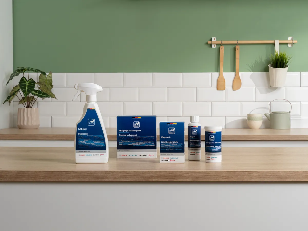 Una collezione di prodotti per la pulizia e la manutenzione su un piano di lavoro in cucina, con una parete piastrellata bianca e una parete verde.