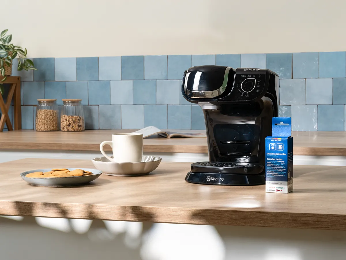 En svart kaffebryggare på en bänkskiva i trä, med en mugg, kakor och en förpackning gräddersättning, mot ett ljusblått kaklat stänkskydd.