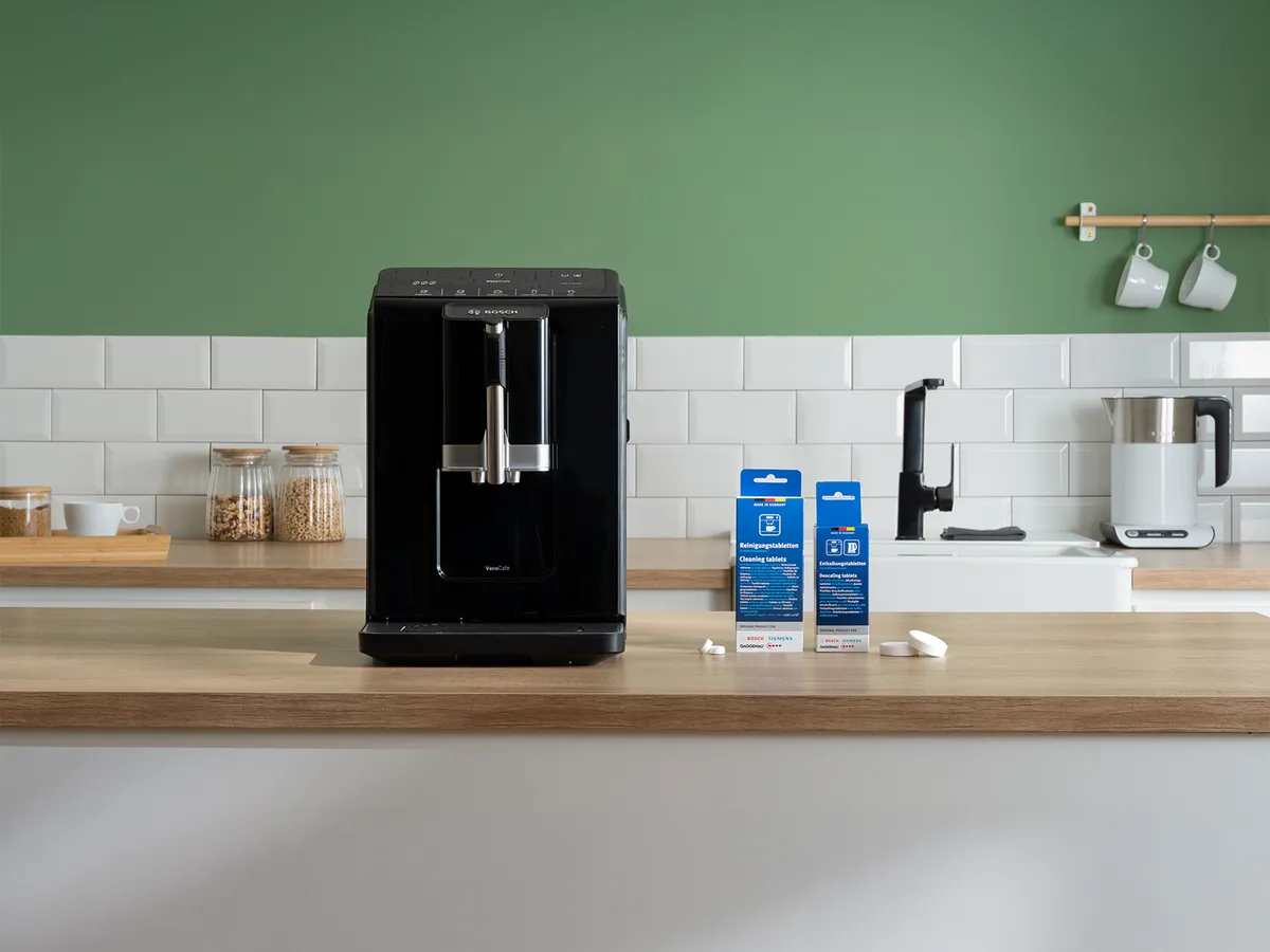 En stilren svart vattendispenser på en köksbänk i trä med rengöringsprodukter bredvid, med en grön vägg i bakgrunden.