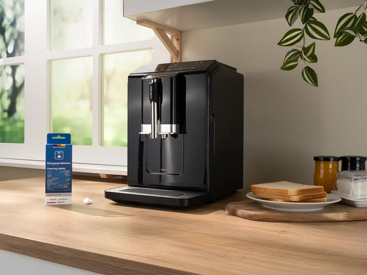 En modern svart kaffemaskin står på en träbänk bredvid en förpackning rengöringstabletter och en tallrik med rostat bröd. Solljuset strömmar in.
