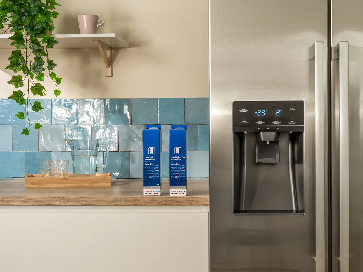 Cuisine moderne avec un frigo en acier inox avec distributeur d’eau, une crédence en carrelage bleu et des verres sur un plateau en bois.