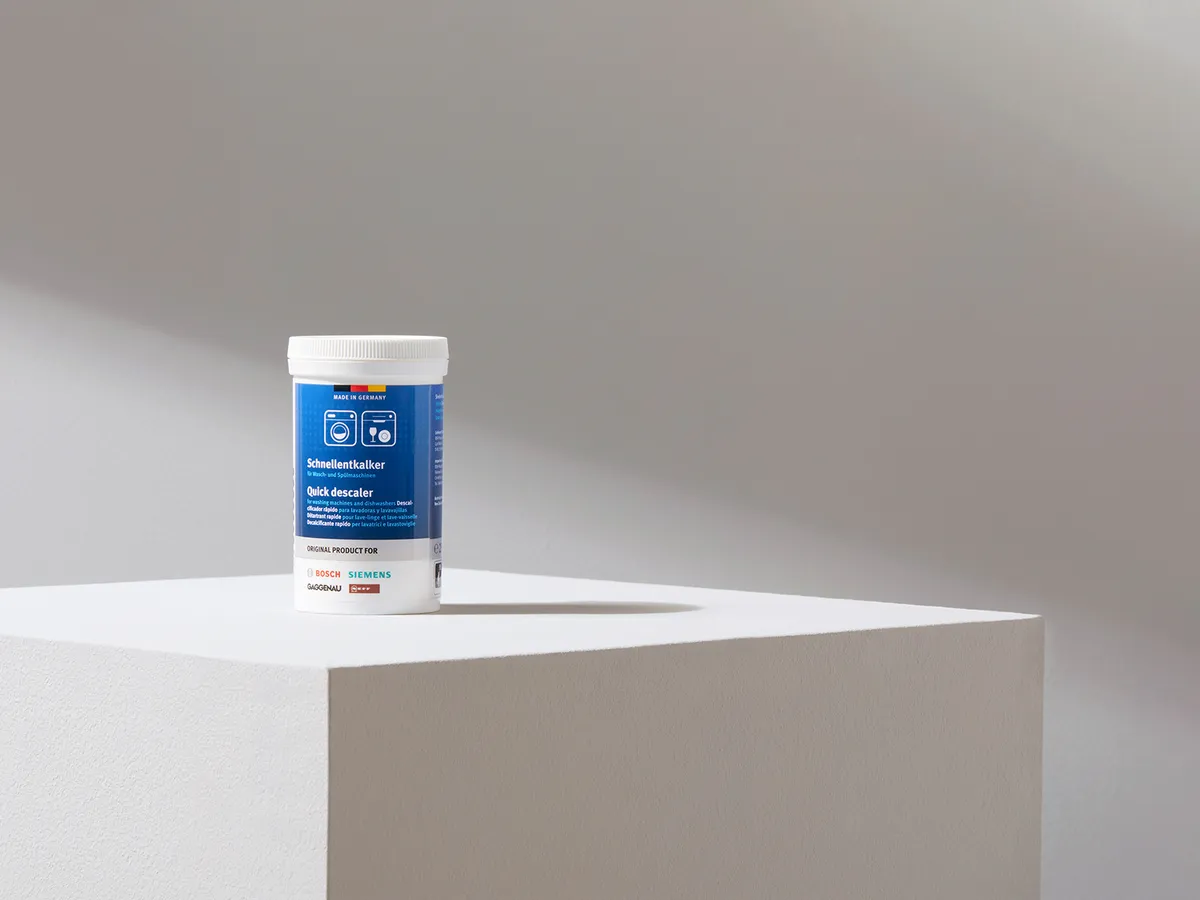 Un bidon de « Quick Descaler » posé sur un socle blanc devant un mur gris clair, portant une étiquette bleue avec des logos d'appareils électroménagers.