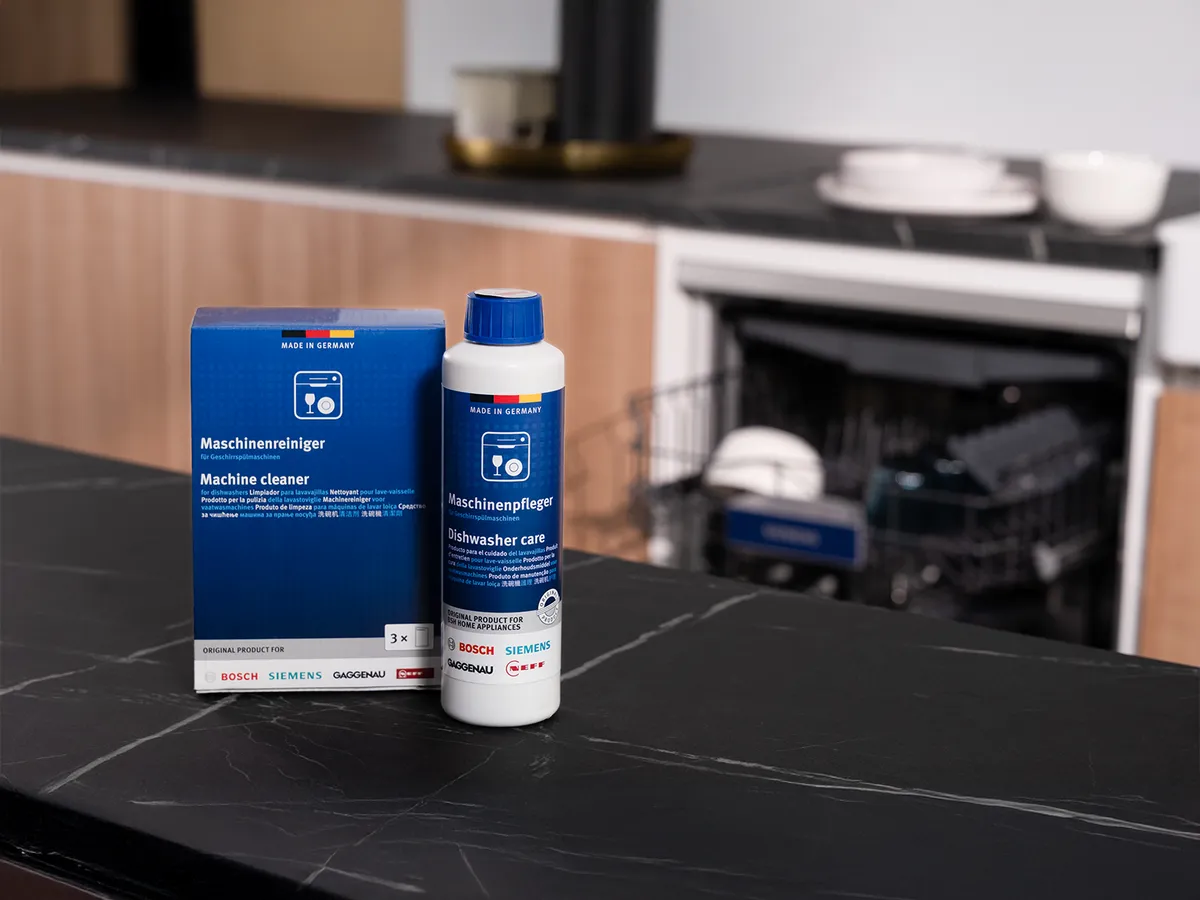Un flacon de produit d'entretien pour lave‑vaisselle et une boîte de nettoyant pour appareil sont posés sur un plan de travail noir, avec un lave‑vaisselle en arrière‑plan.