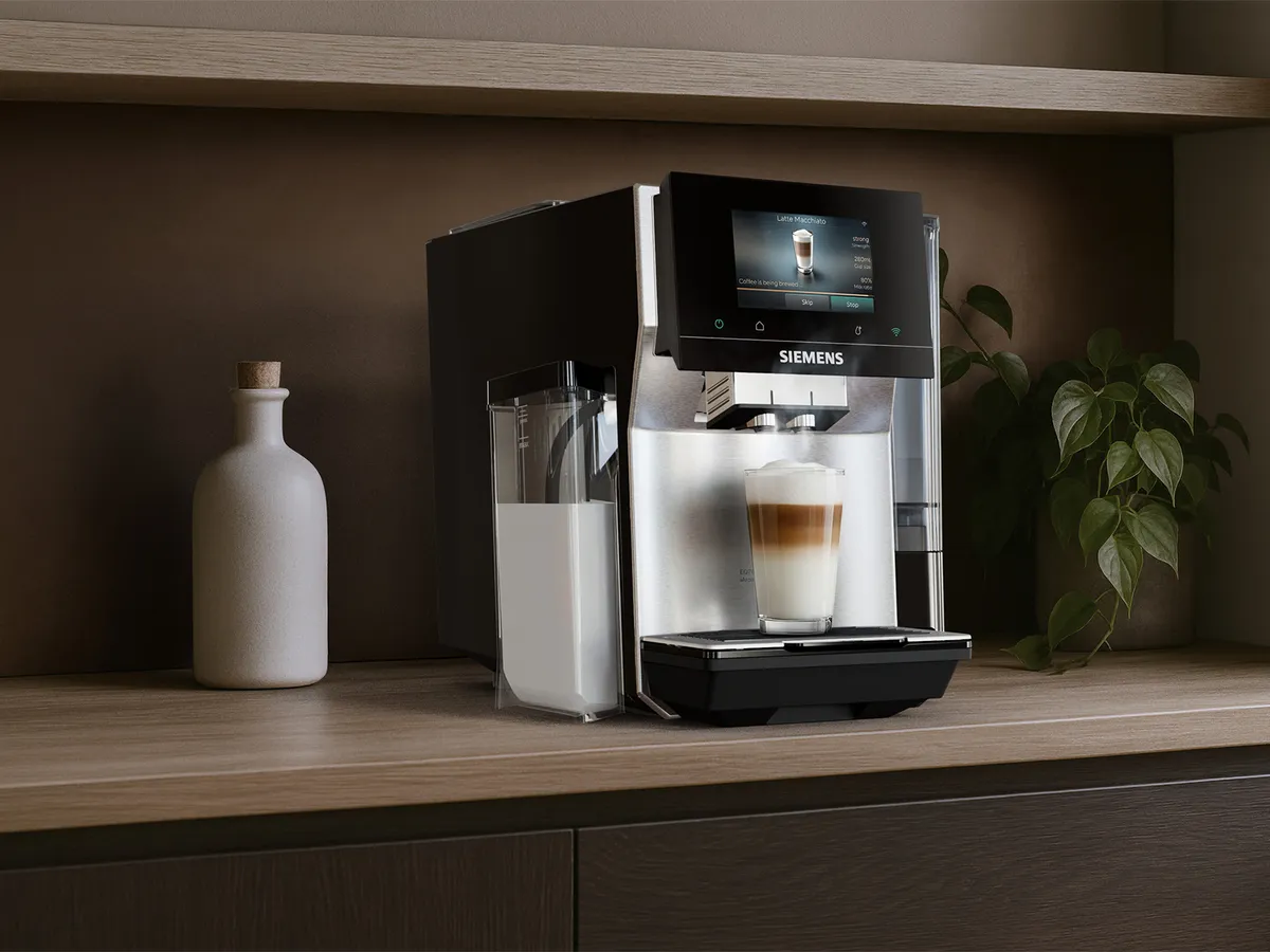 Siemens coffee machine with attached milk container on a wooden countertop with a layered latte macchiato; small beige vase with wooden lid and small plant in vase.