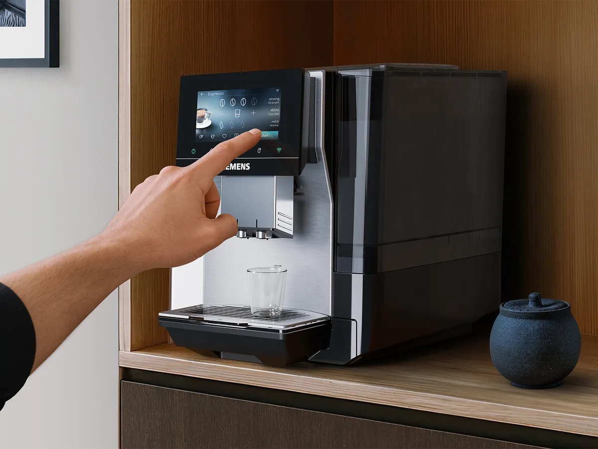 Male hand is selecting his personal preferences on the touch screen display of a Siemens coffee machine which stands on a wooden countertop with a black sugar jar next to the machine.