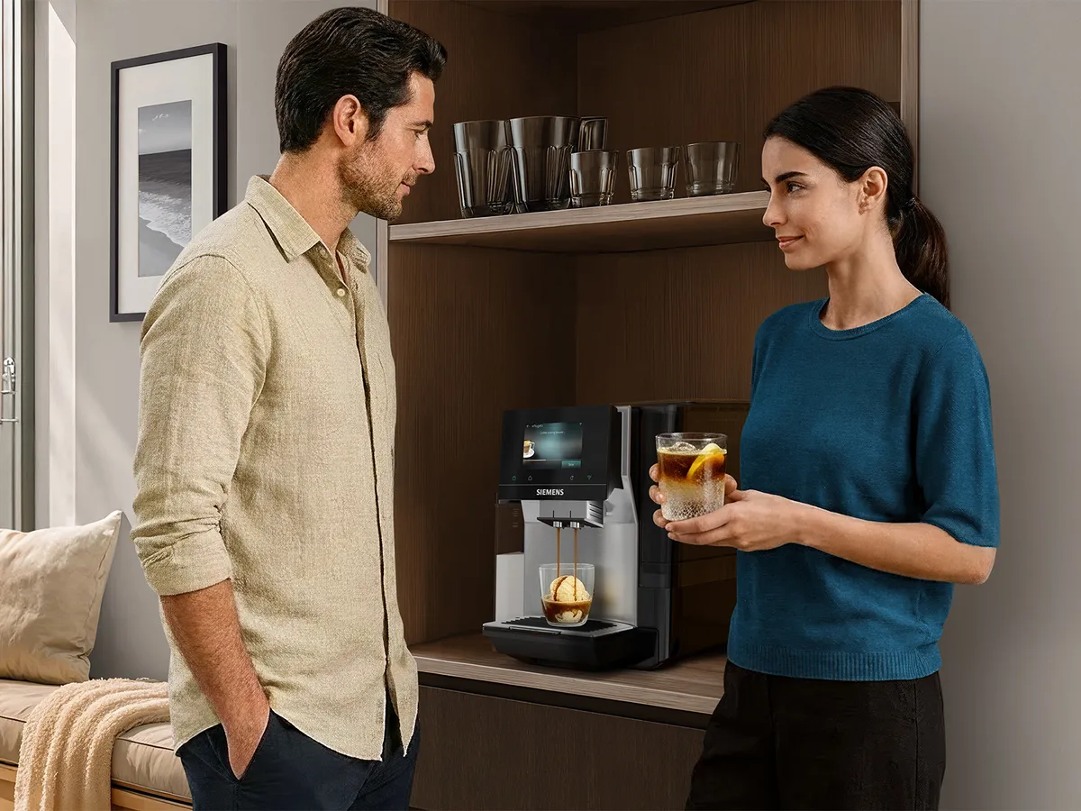 A man and woman are talking in a bright kitchen while a Siemens coffee machine brews an espresso into a glass with vanilla ice cream.