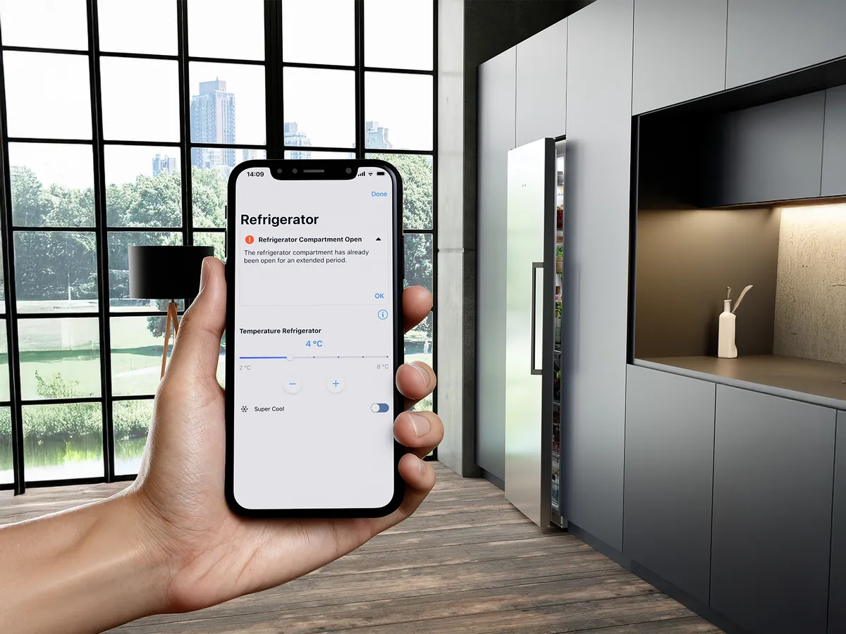 A hand holds a smartphone displaying a refrigerator app warning of an open compartment, with a modern kitchen in the background.