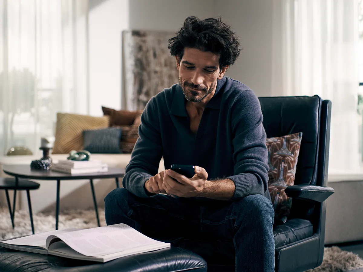 Person sitting in an armchair using a smartphone, open book on ottoman in a cozy living room.