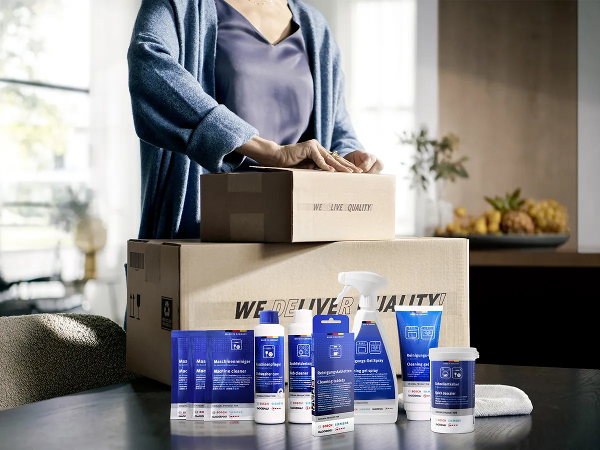 A person in a blue cardigan packs cleaning supplies and a box labeled “WE DELIVER QUALITY” on a kitchen table.