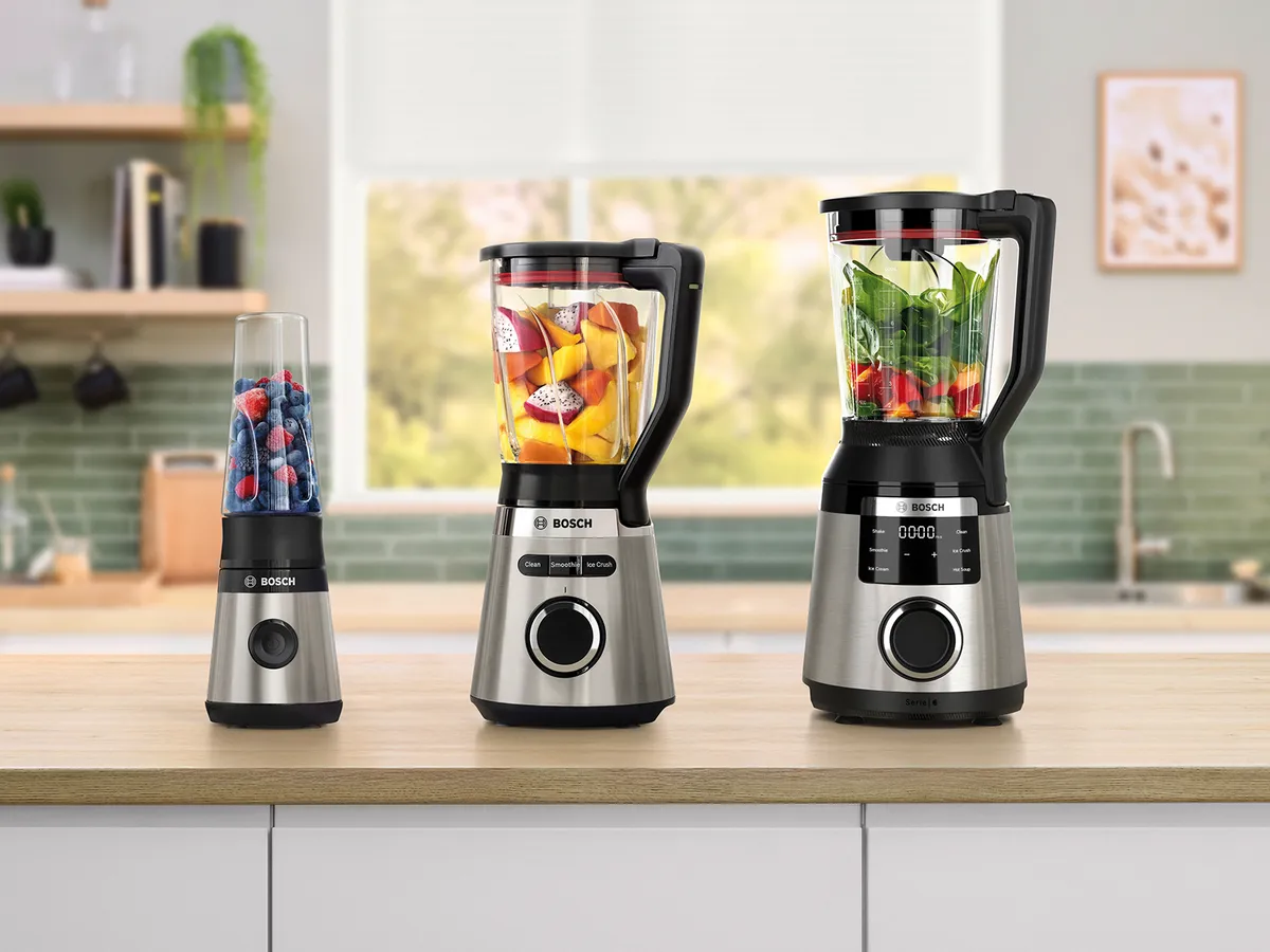 Drei Bosch-Standmixer mit Obst und Gemüse im Inneren stehen auf einer Küchenarbeitsplatte vor einem unscharfen Fenster im Hintergrund.