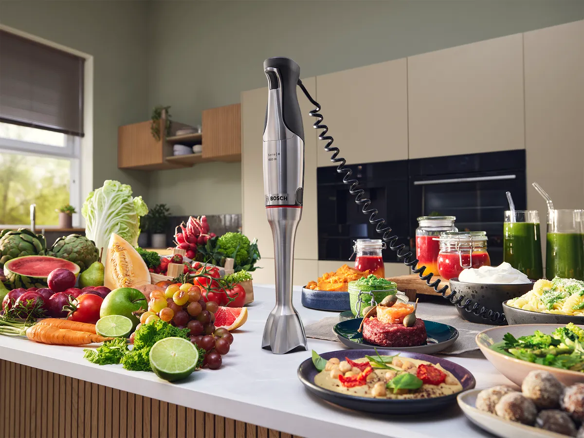 Bosch Stabmixer steht auf einer Küchentheke, umgeben von frischem Obst, Gemüse und leckeren Gerichten.