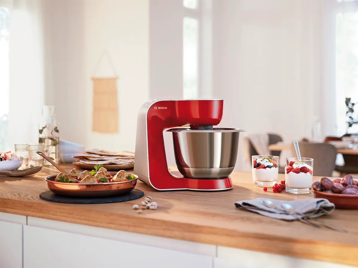 Eine leuchtend rote Küchenmaschine steht auf einer Holzarbeitsplatte in der Küche, umgeben von zubereiteten Speisen, darunter Desserts und Snacks, in einer hellen, modernen Umgebung.