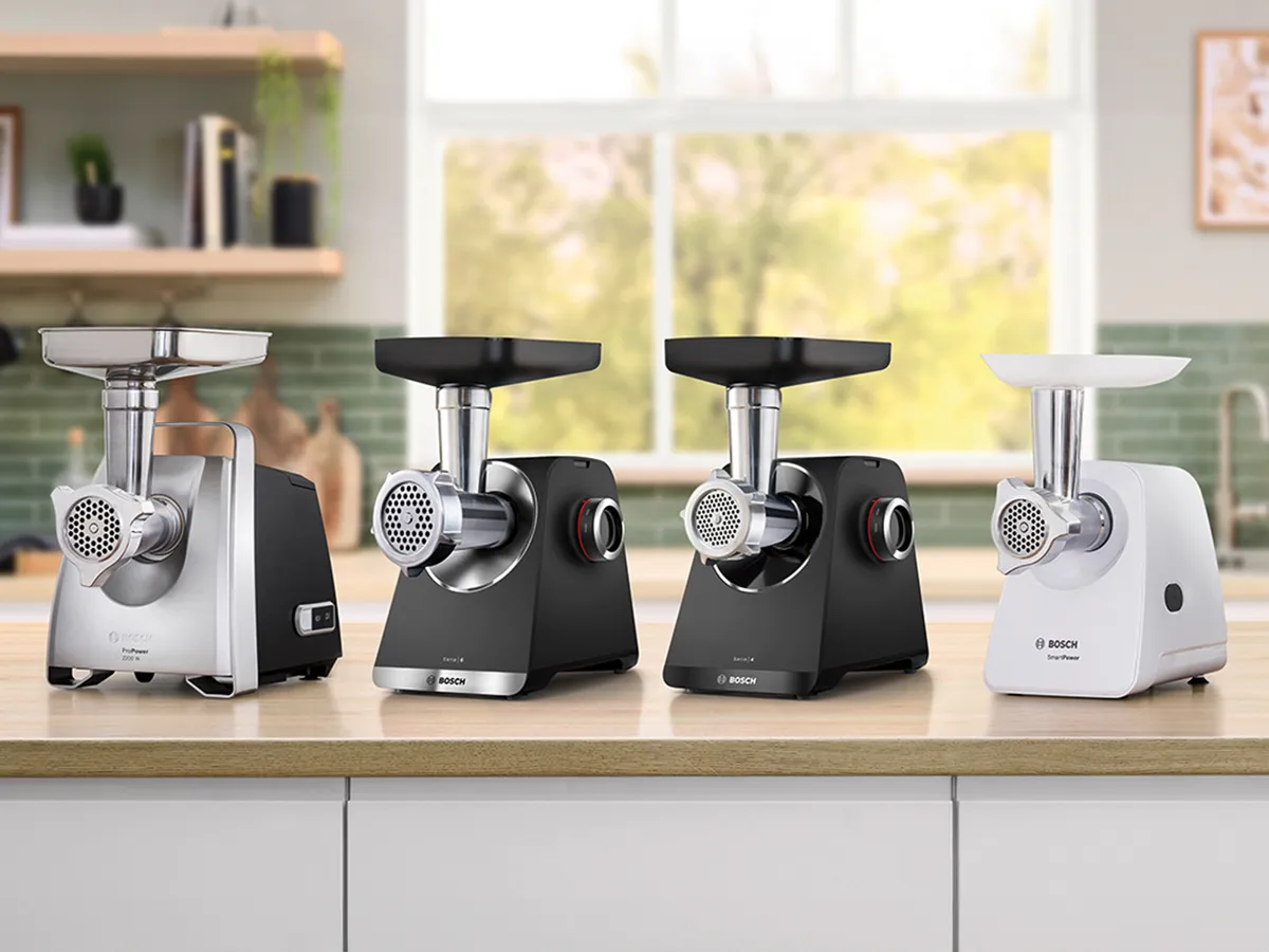 Four Bosch meat grinders in silver, black, and white, displayed on a kitchen countertop with a green tiled background.