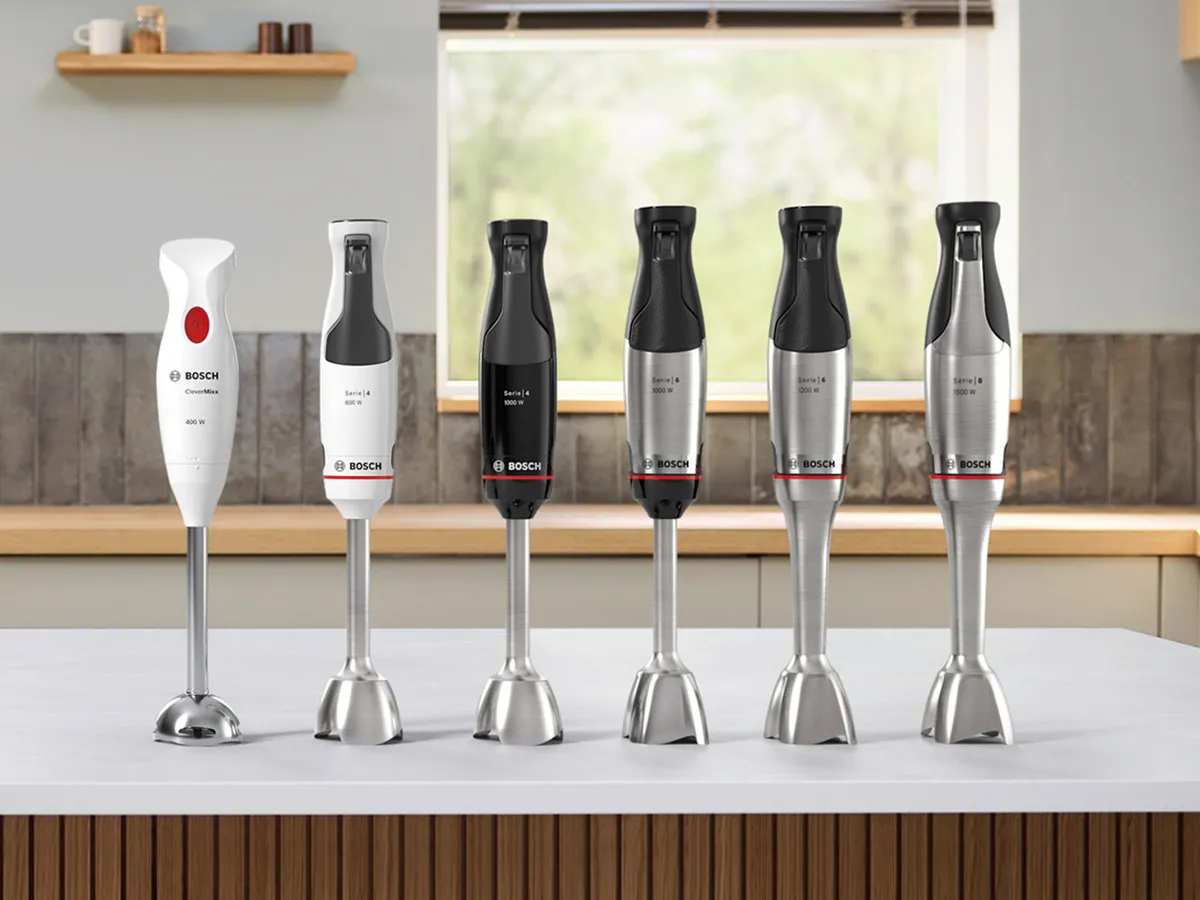 Sechs Bosch Stabmixer in verschiedenen Modellen und Farben auf weißer Küchenarbeitsplatte vor einem Fenster.