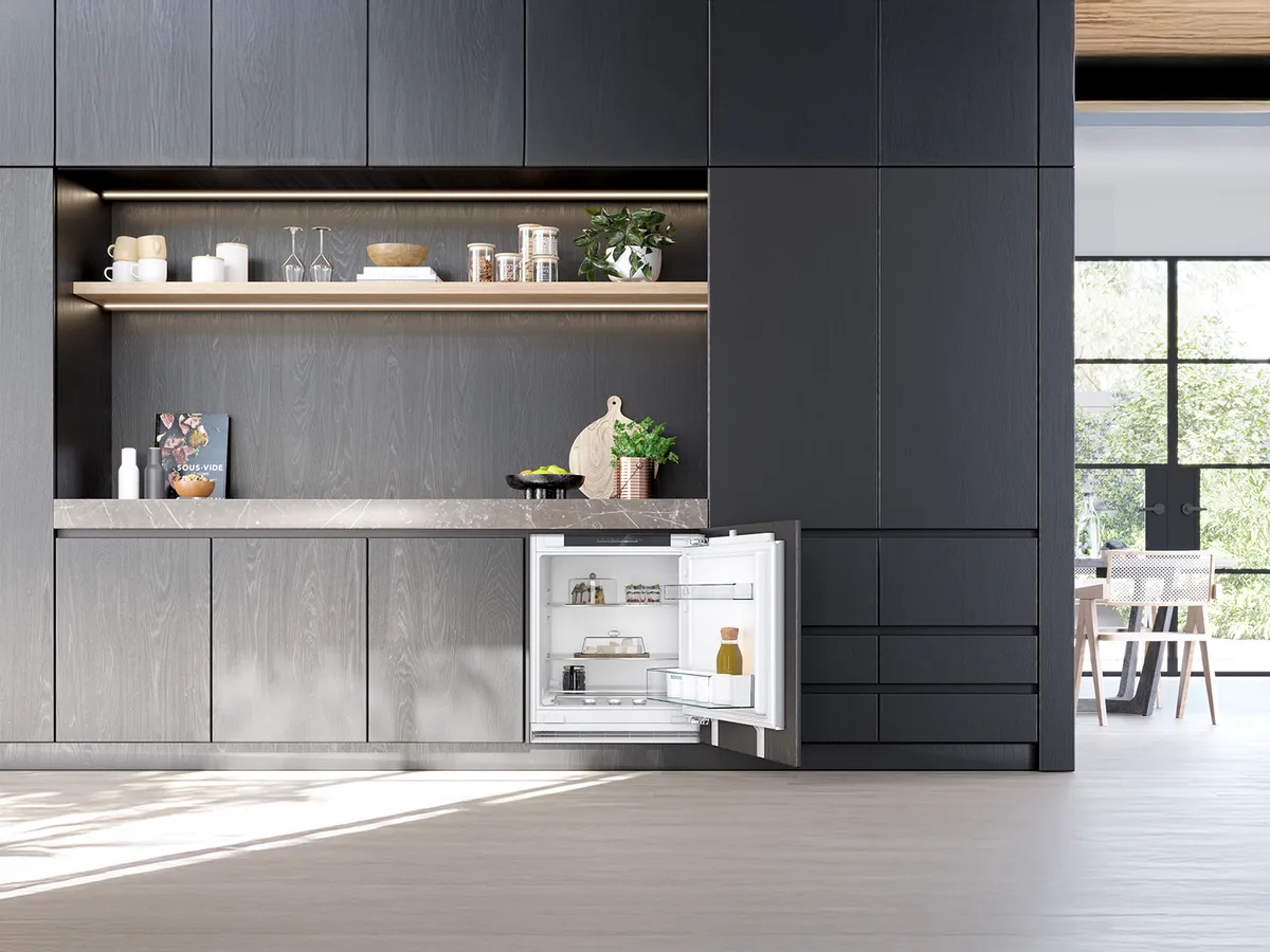 A sleek, modern kitchen with dark cabinetry, an open under-counter refrigerator, and a view into a dining area.
