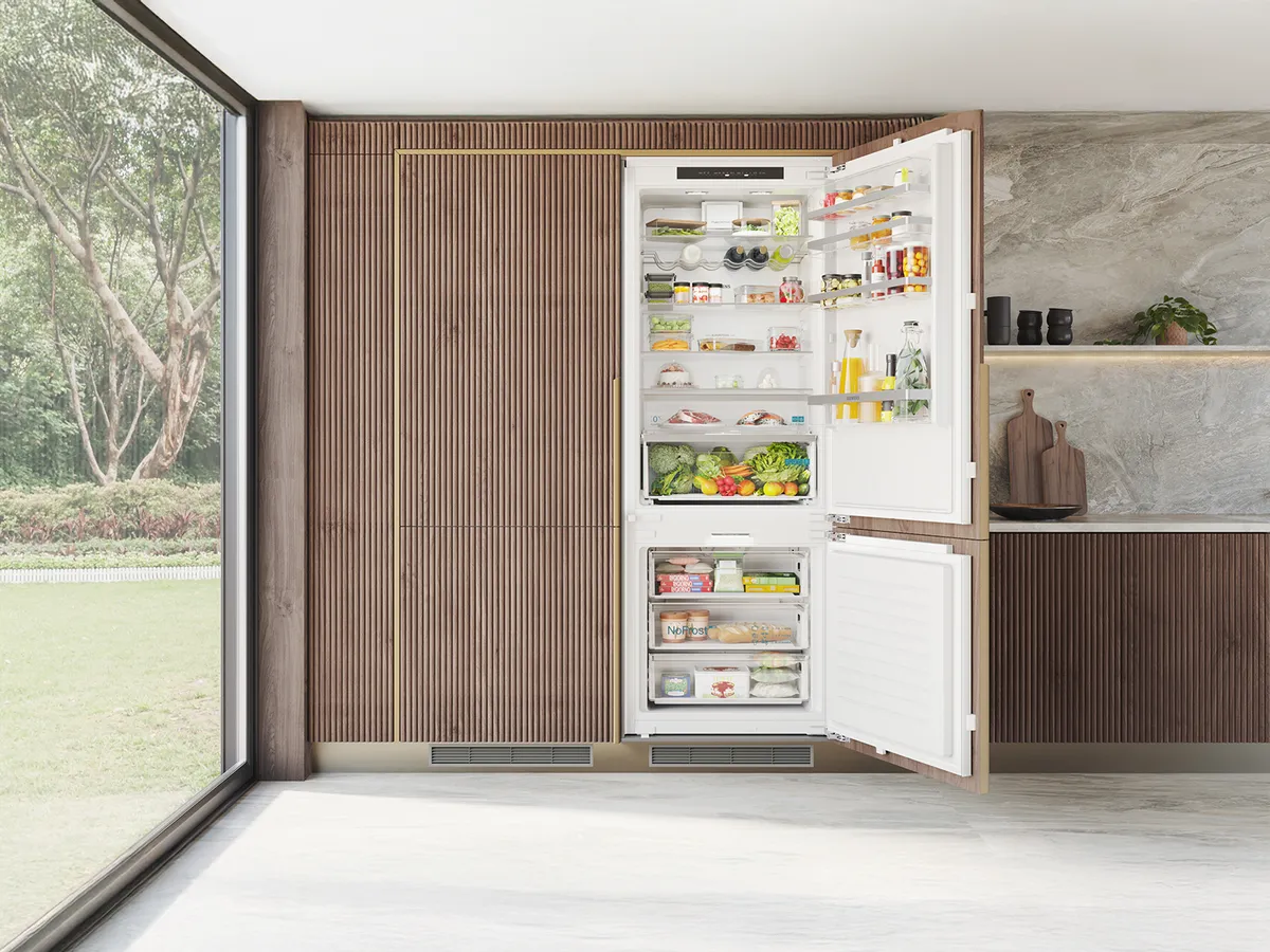 A modern kitchen with a large, open refrigerator filled with food, next to a window overlooking a garden.