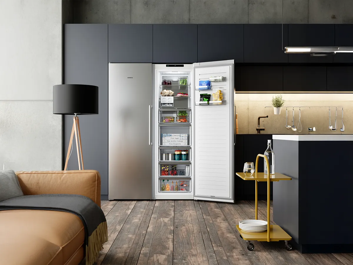 Modern kitchen featuring an open fridge filled with food, a cozy sofa, stylish lamp, and a yellow cart with glassware and dishes.