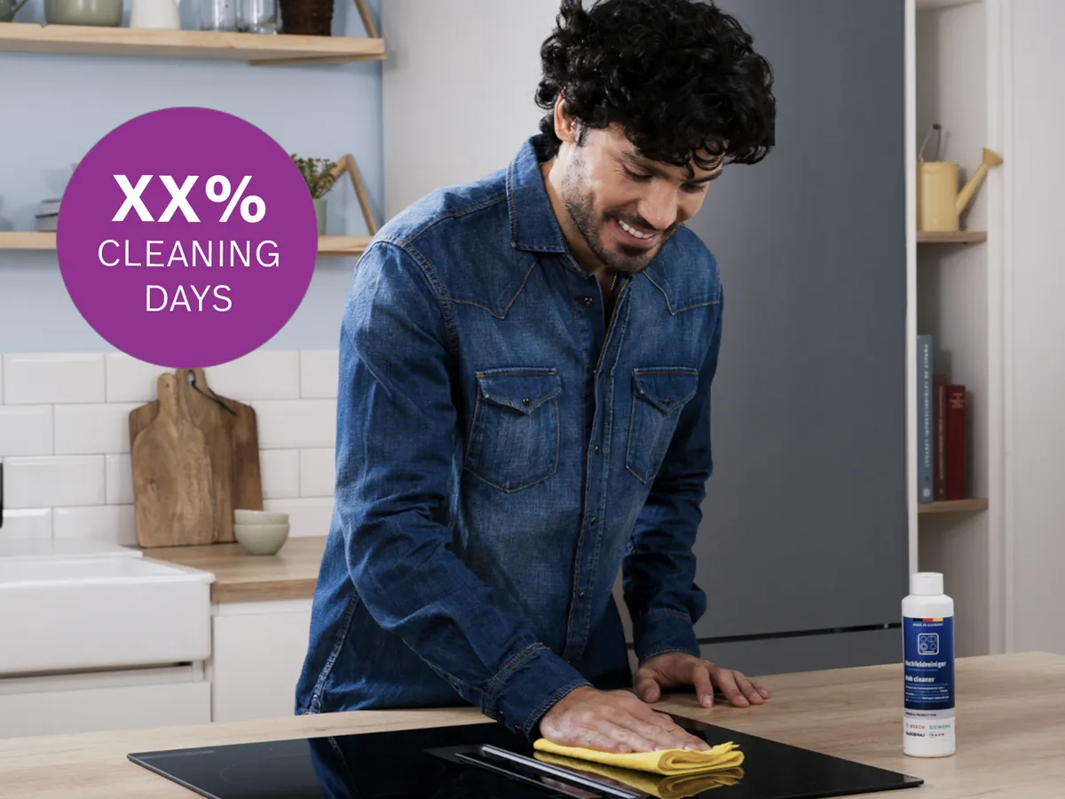  Person wiping induction cooktop with yellow cloth, Bosch cleaning bottle on counter, purple "XX% CLEANING DAYS" badge top-left.