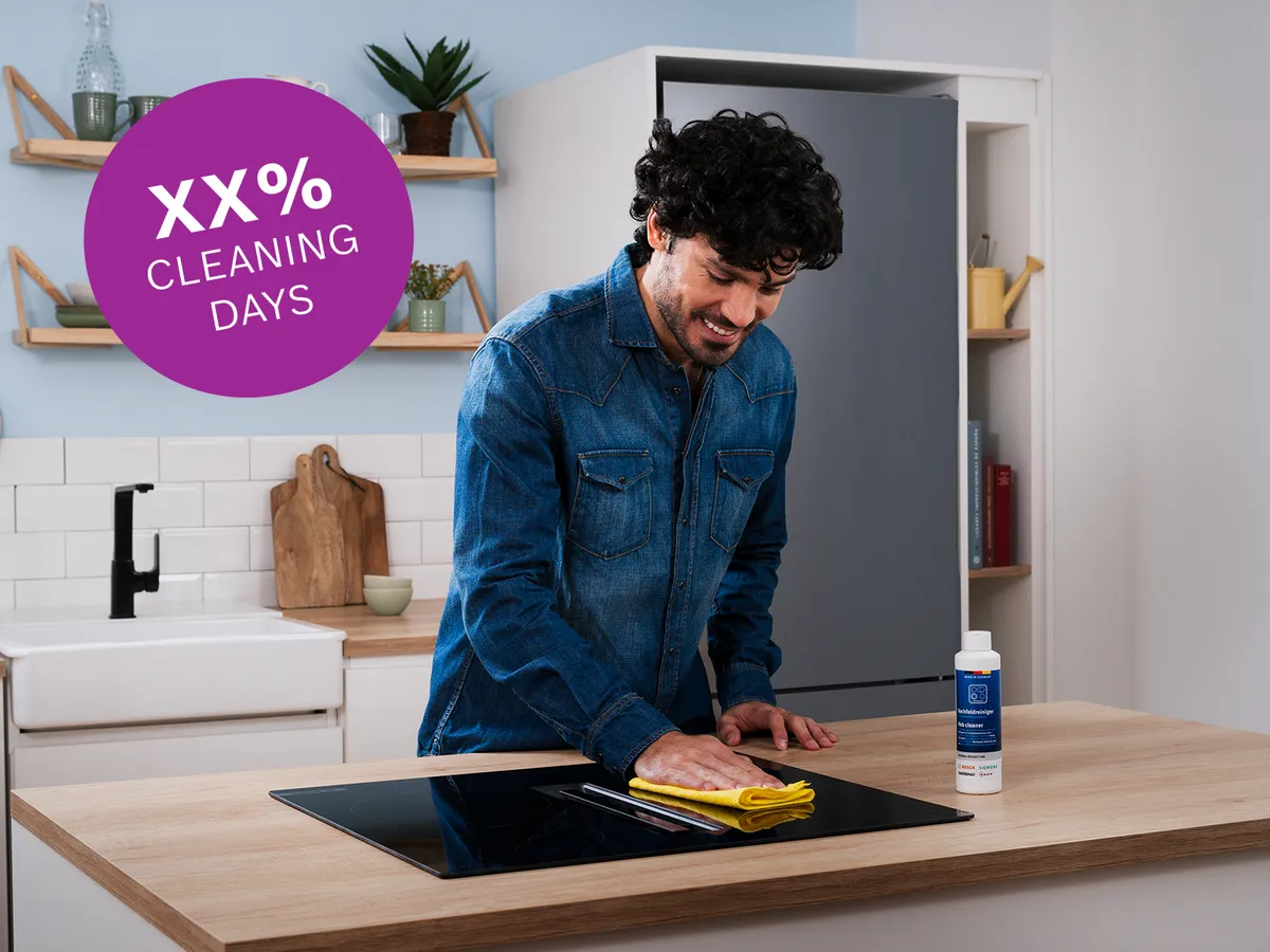 Person wiping induction cooktop with yellow cloth, Bosch cleaning bottle on counter, purple "XX% CLEANING DAYS" badge top-left.