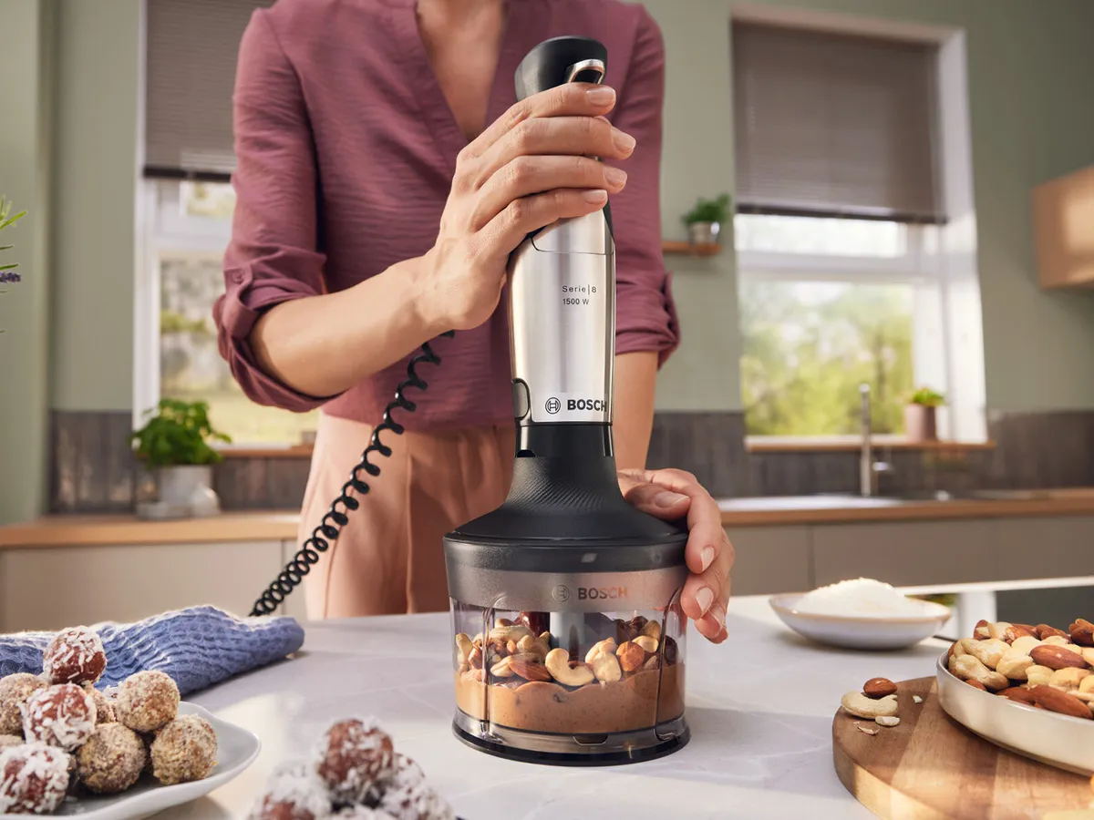 Una batidora de mano MaxoMixx de Bosch descansa junto a boles de salsa de naranja y puré verde sobre una superficie clara, mostrando su elegante diseño.