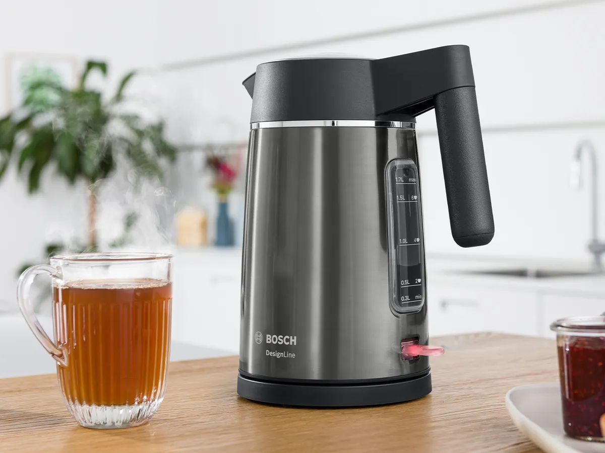 Bosch DesignLine Wasserkocher mit Wasserstandsanzeige und schwarzem Griff auf einer Holzküchenarbeitsplatte – daneben steht ein gefülltes dampfendes Teeglas.