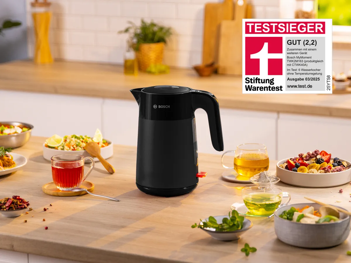 Schwarzer Bosch MyMoment Wasserkocher auf Küchenholzarbeitsplatte, umgeben von Tassen und kleinen Speisen, mit Testsieger‑Logo (Stiftung Warentest, GUT 2,2).