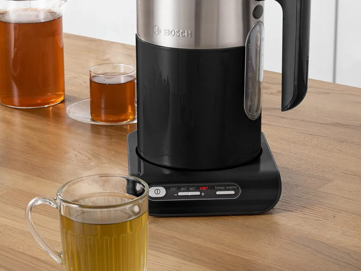 Ein Bosch Wasserkocher steht auf seiner Basisstation, im Vordergrund ein Glas grüner Tee, im Hintergrund mehrere Teegläser auf einem Holztisch.