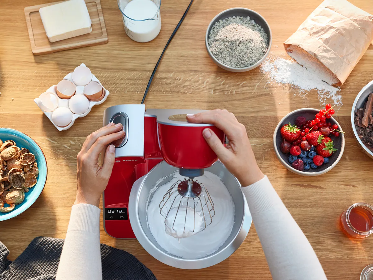 Eine Person verwendet eine rote Küchenmaschine, um Zutaten zu vermischen, umgeben von Eiern, Mehl, Beeren und verschiedenen Backzutaten auf einer Holzarbeitsplatte.