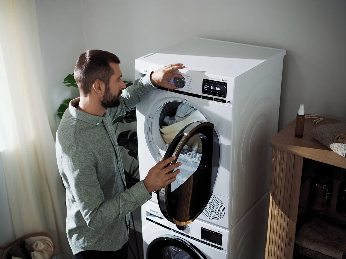 Ein Mann öffnet die Tür eines weißen, übereinander gestapelten Trockners und stellt die Einstellungen ein. Wäsche ist zu sehen. Die Waschküche ist hell und enthält eine passende Waschmaschine.