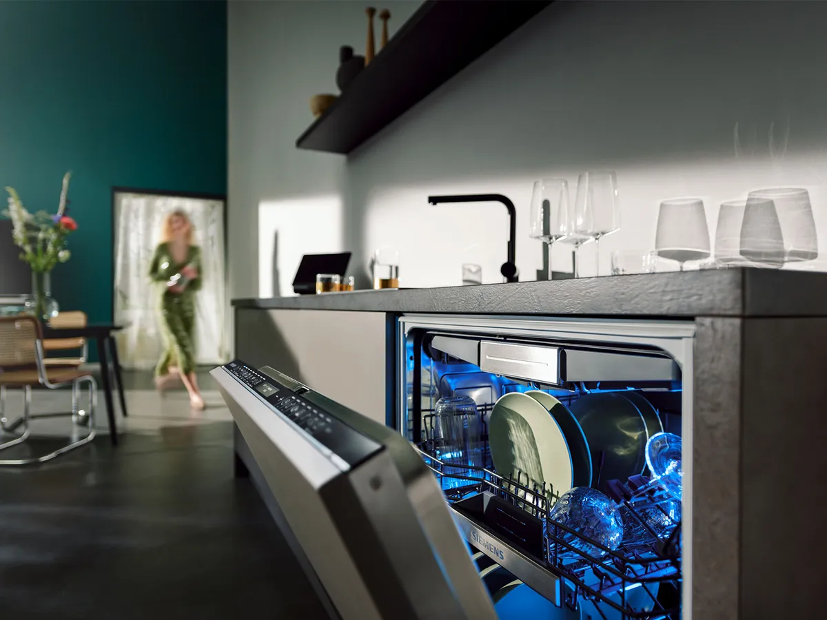 Eine moderne Küchenszene zeigt einen geöffneten Geschirrspüler mit blau leuchtendem Innenraum, in dem sauberes Geschirr und Gläser zu sehen sind, während eine Person den Raum betritt.