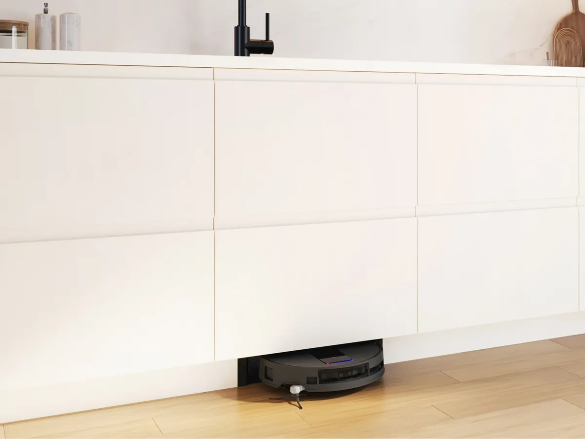 A dark gray robot vacuum cleaner is partially visible, neatly docked or charging in a custom recess under sleek, handleless white kitchen cabinets. The modern kitchen features a black faucet on a white countertop and light wood flooring.
