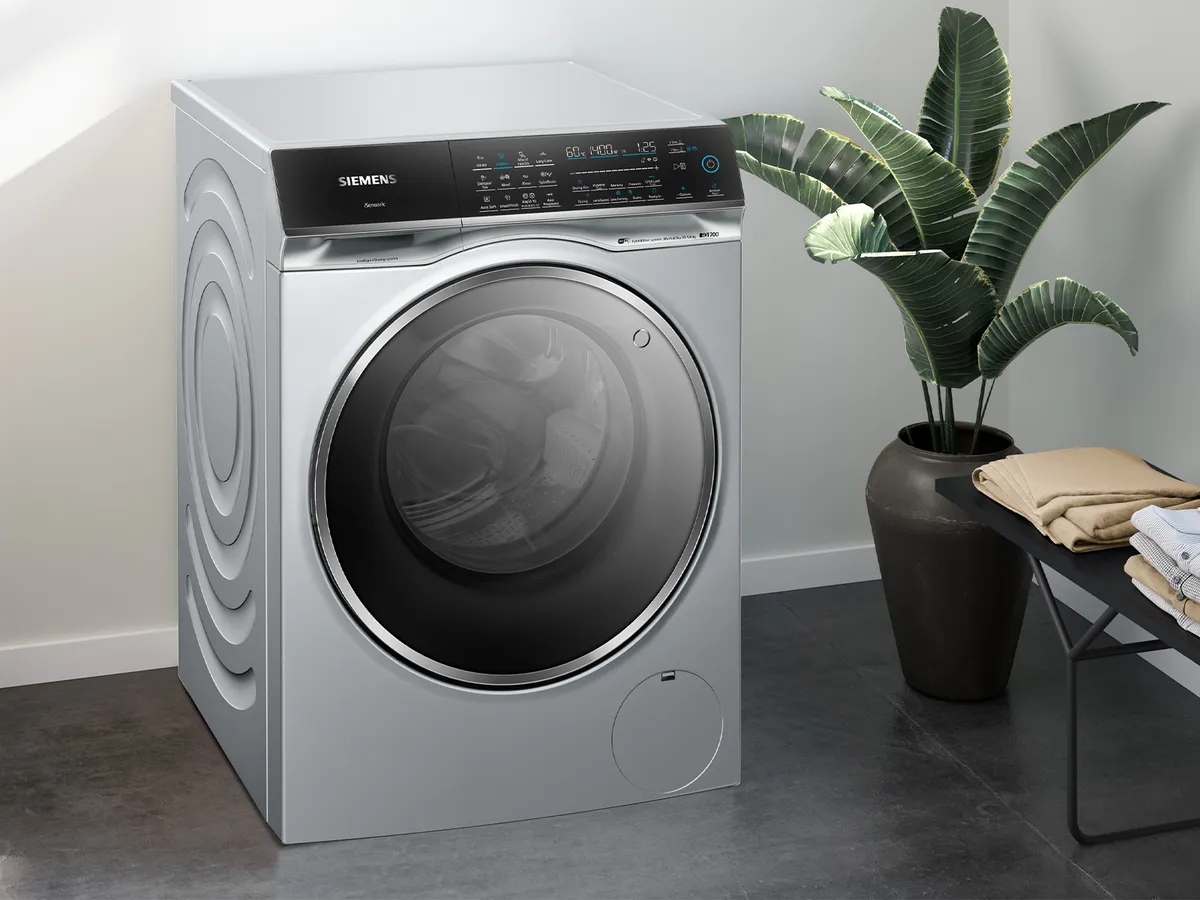 White Siemens washing machine in a sunlit room, beside laundry basket and detergent bottles.