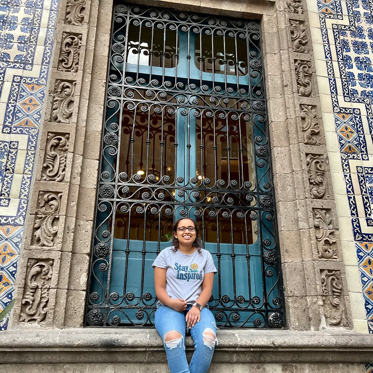 Smyrna’s wearing a "Stay Inspired" shirt and ripped jeans sitting on a stone ledge in front of an ornate wrought iron window with blue patterned tile walls.