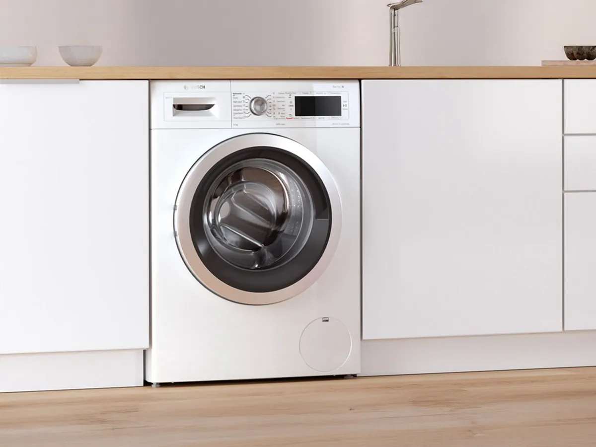 Front-load washer integrated into sleek white kitchen cabinetry with wooden countertop and minimalist accessories.