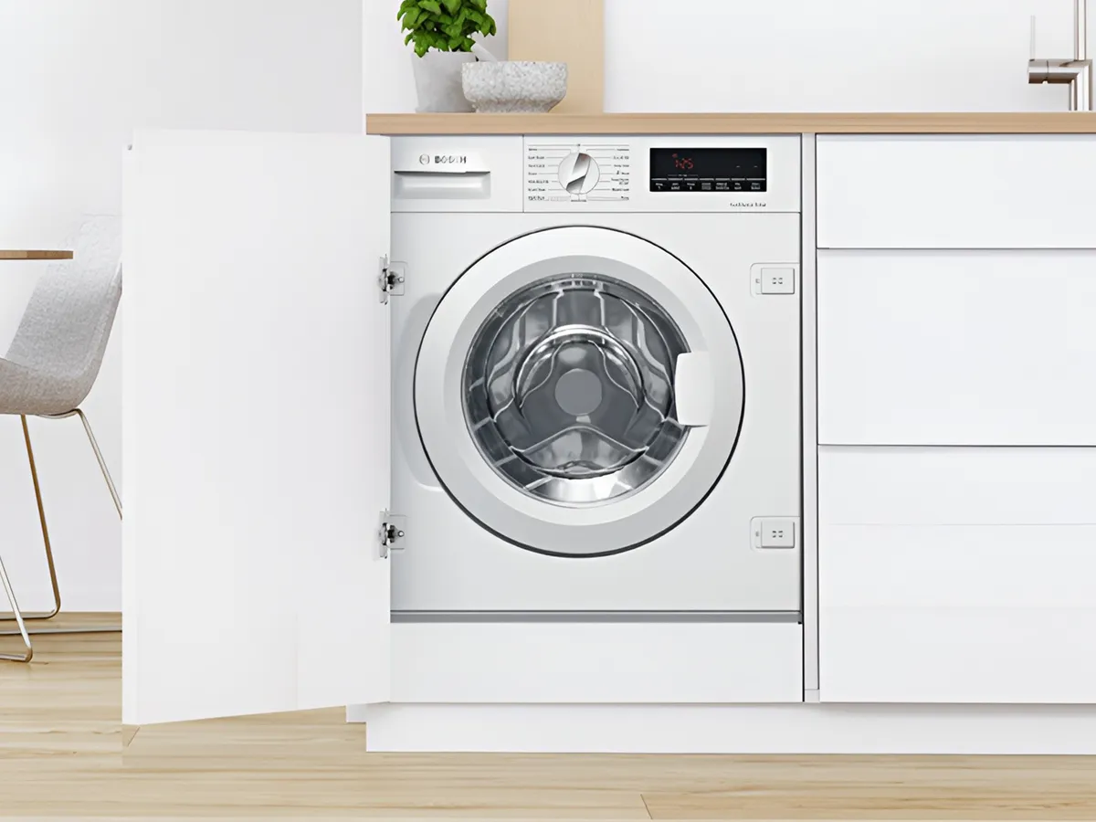 Бяла пералня Bosch вградена в модерен кухненски шкаф, с леко открехната врата.