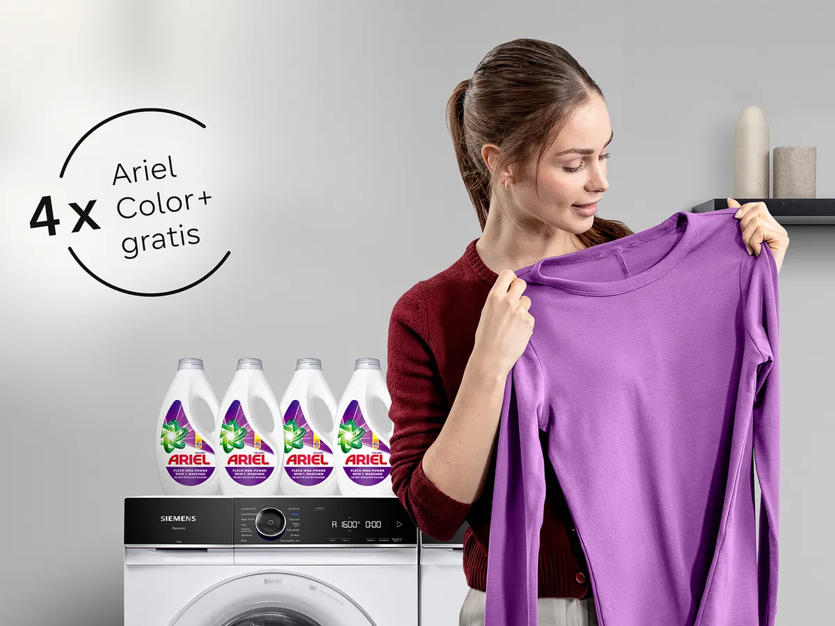 Eine Frau hält einen Lila Pullover hoch und steht vor einer Siemens Waschmaschine. Auf der Waschmaschine stehen 4 Flaschen Ariel Color Waschmittel.
