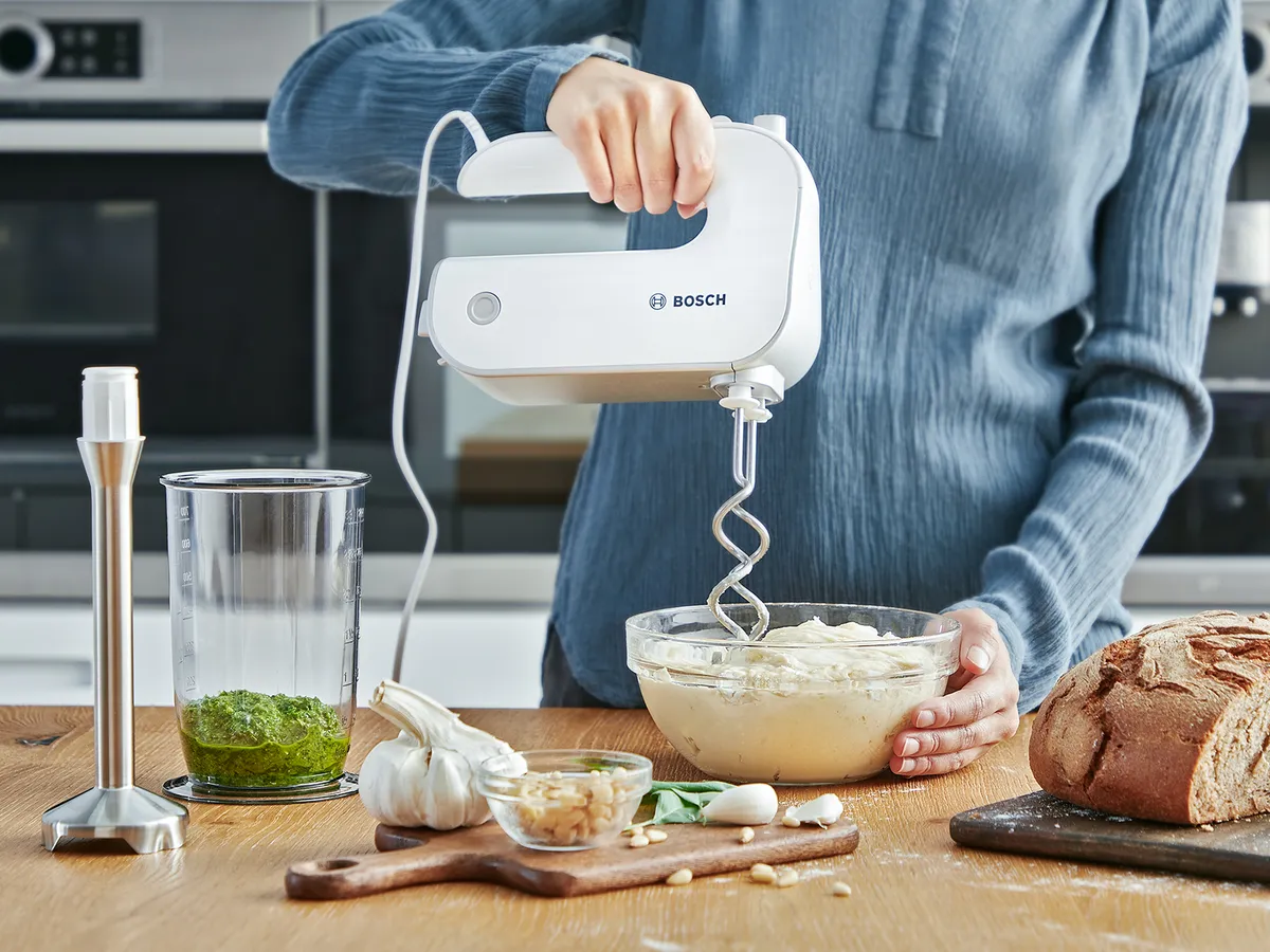 Person hält Bosch-Handmixer über Schüssel mit Teig auf Holztisch mit Brot, Knoblauch und Kräutern