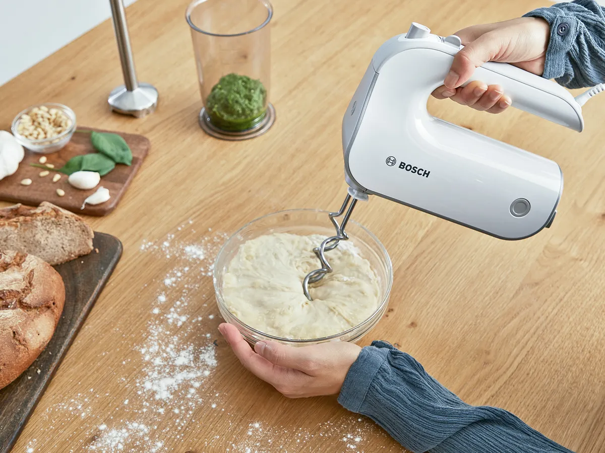 Hand hält weißen Bosch-Handmixer über Glasschüssel mit Teig auf Holzarbeitsfläche mit Mehl, Brot und Kräutern