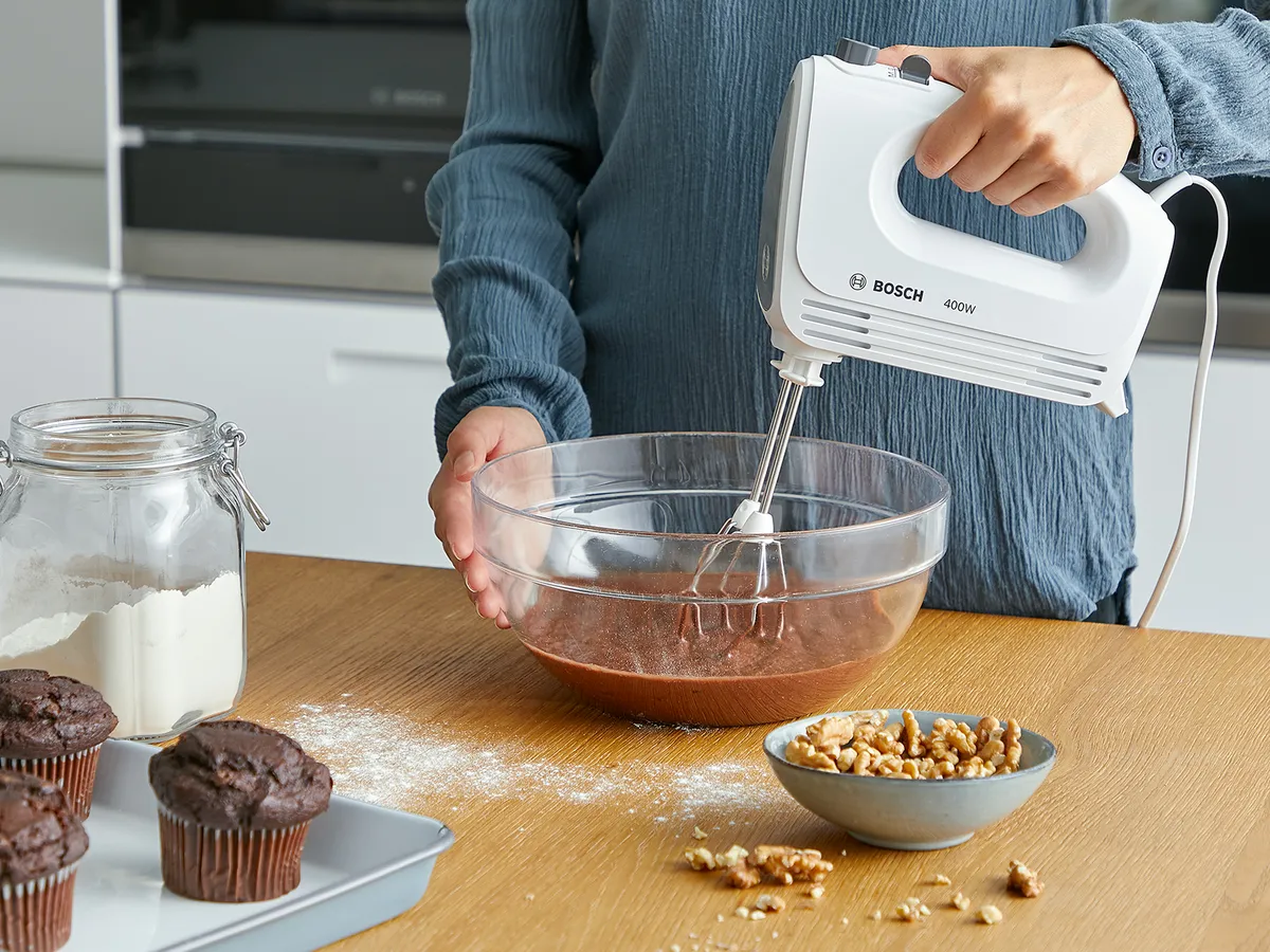 Eine Person verwendet einen Bosch-Handmixer, um Schokoladenteig in einer Glasschüssel auf einer Küchenarbeitsplatte zu verrühren; in der Nähe befinden sich Muffins, Mehl und Walnüsse.