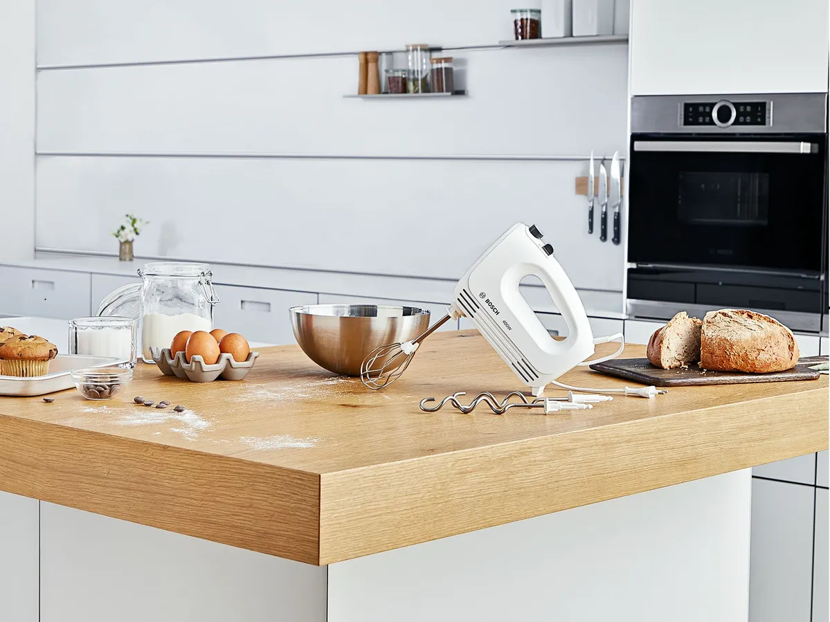 Moderne Küchenarbeitsplatte mit Backzutaten, einem Handmixer, Eiern, einer Schüssel und frisch gebackenem Brot auf einem Schneidebrett.