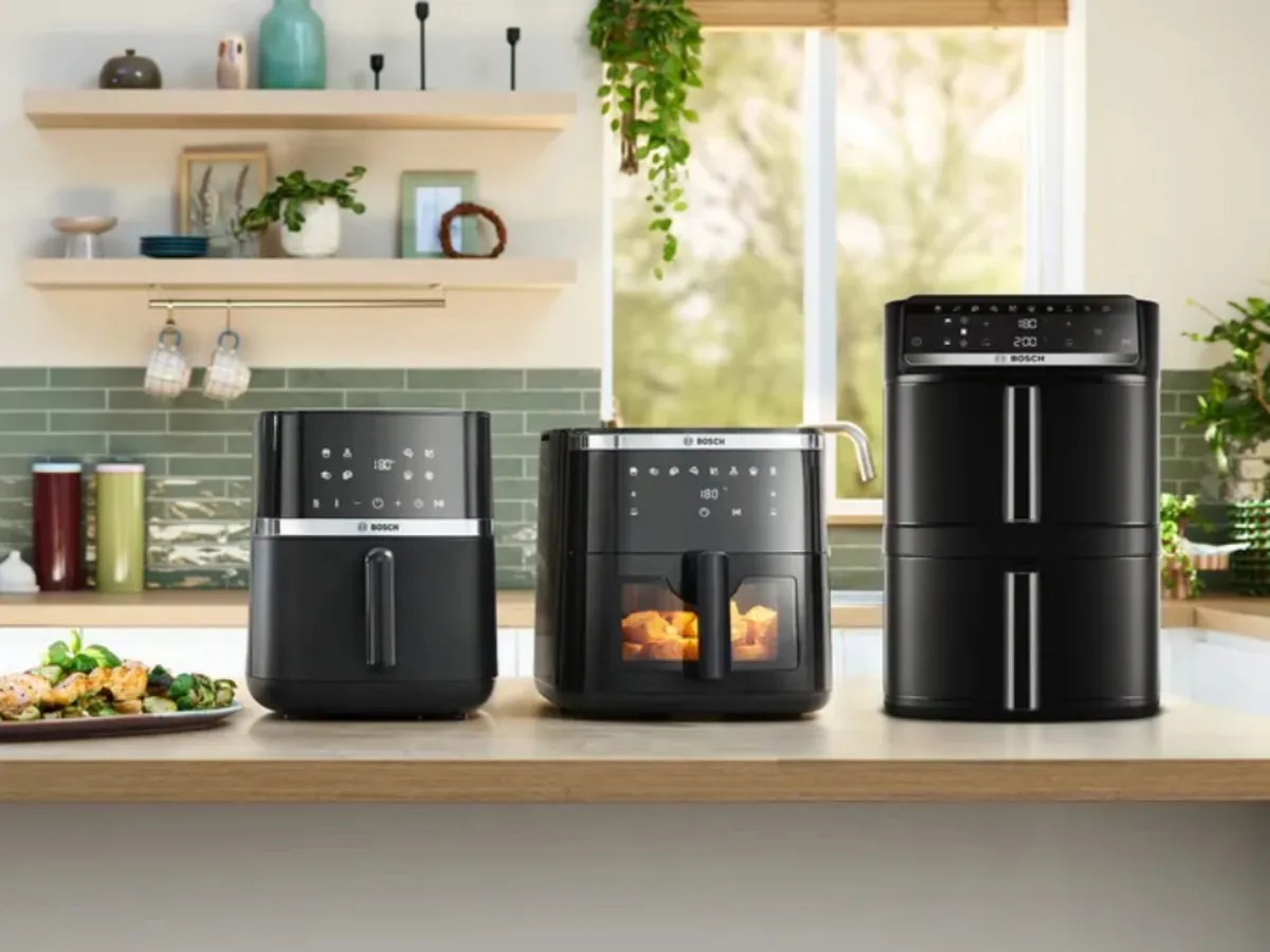 Moderne Küchenarbeitsplatte mit einer Heißluftfritteuse, einem Mini-Backofen und einem Multikocher, sowie zubereiteten Gerichten und Grünpflanzen vor hellem Hintergrund.