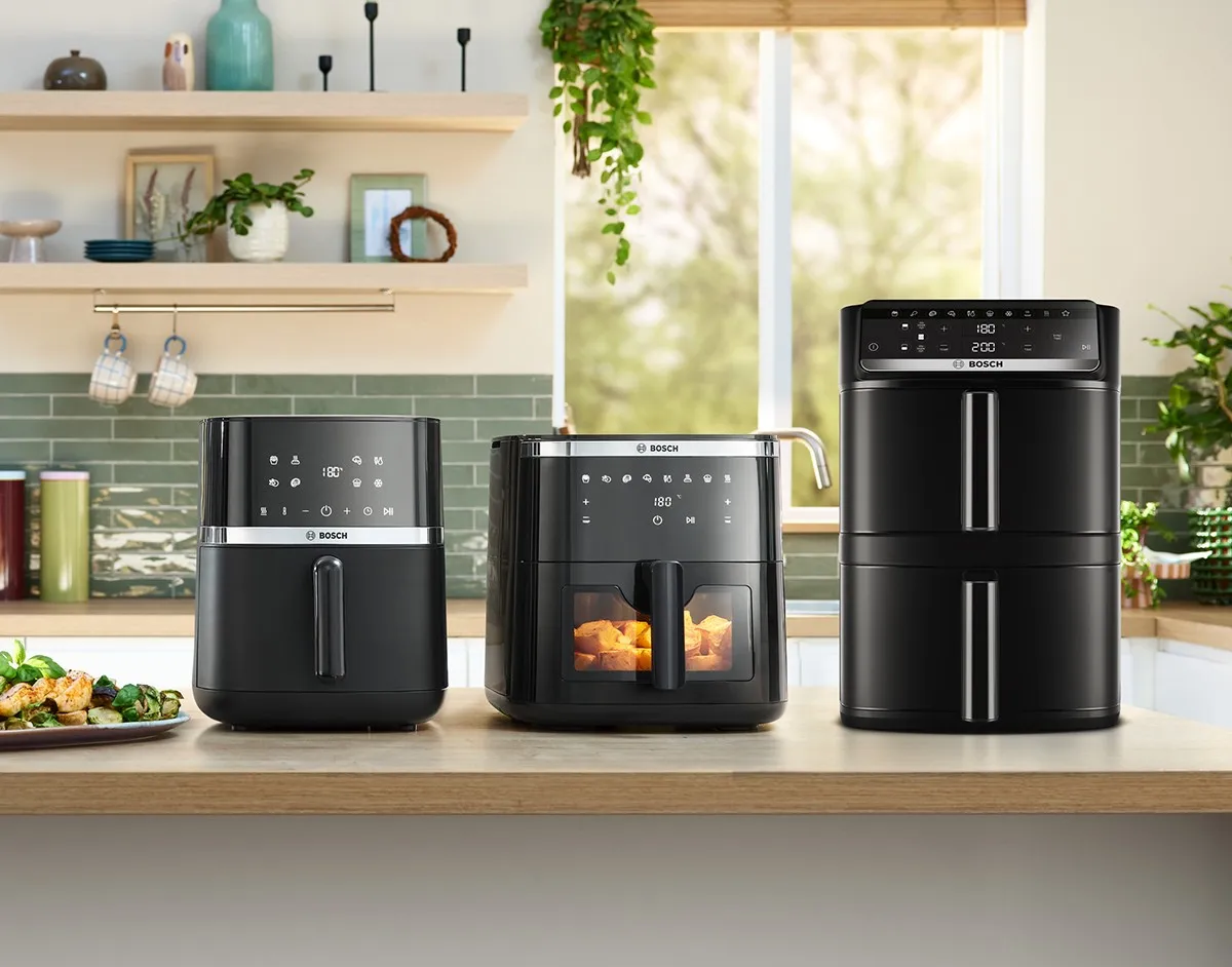 Moderne Küchenarbeitsplatte mit einer Heißluftfritteuse, einem Mini-Backofen und einem Multikocher, sowie zubereiteten Gerichten und Grünpflanzen vor hellem Hintergrund.