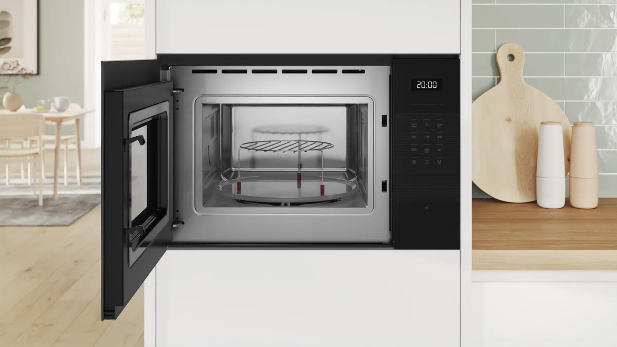 Open built-in microwave with stainless-steel interior, glass turntable and raised metal rack, digital control panel showing "20:00," set in white cabinetry on a light-wood countertop with a round cutting board and pale green tiled backsplash.